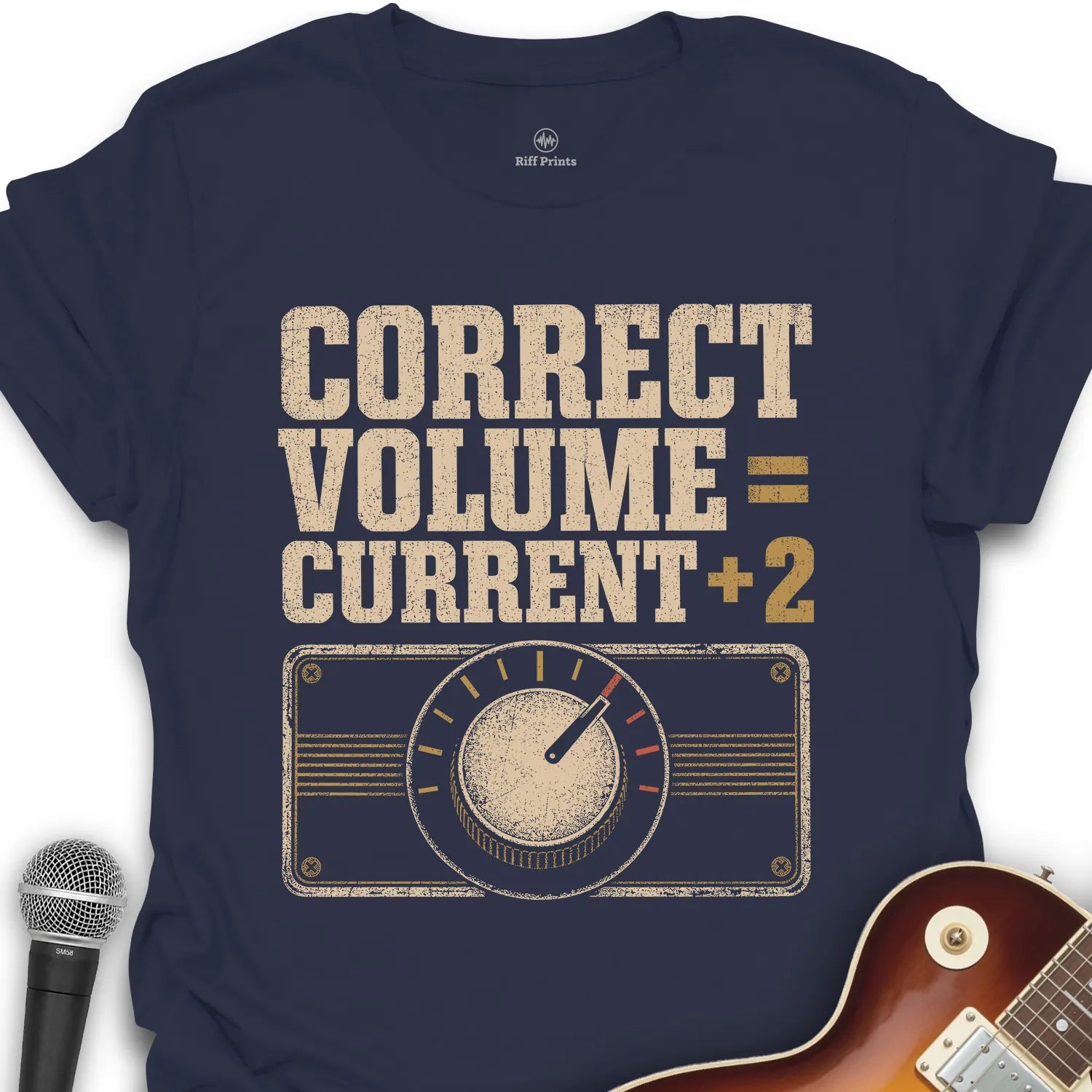 A navy blue graphic t-shirt featuring a retro-style radio dial design and the text 'CORRECT VOLUME = CURRENT + 2' in distressed beige and gold lettering. The t-shirt is displayed flat with a microphone on the left and the corner of a sunburst electric guitar on the bottom right.